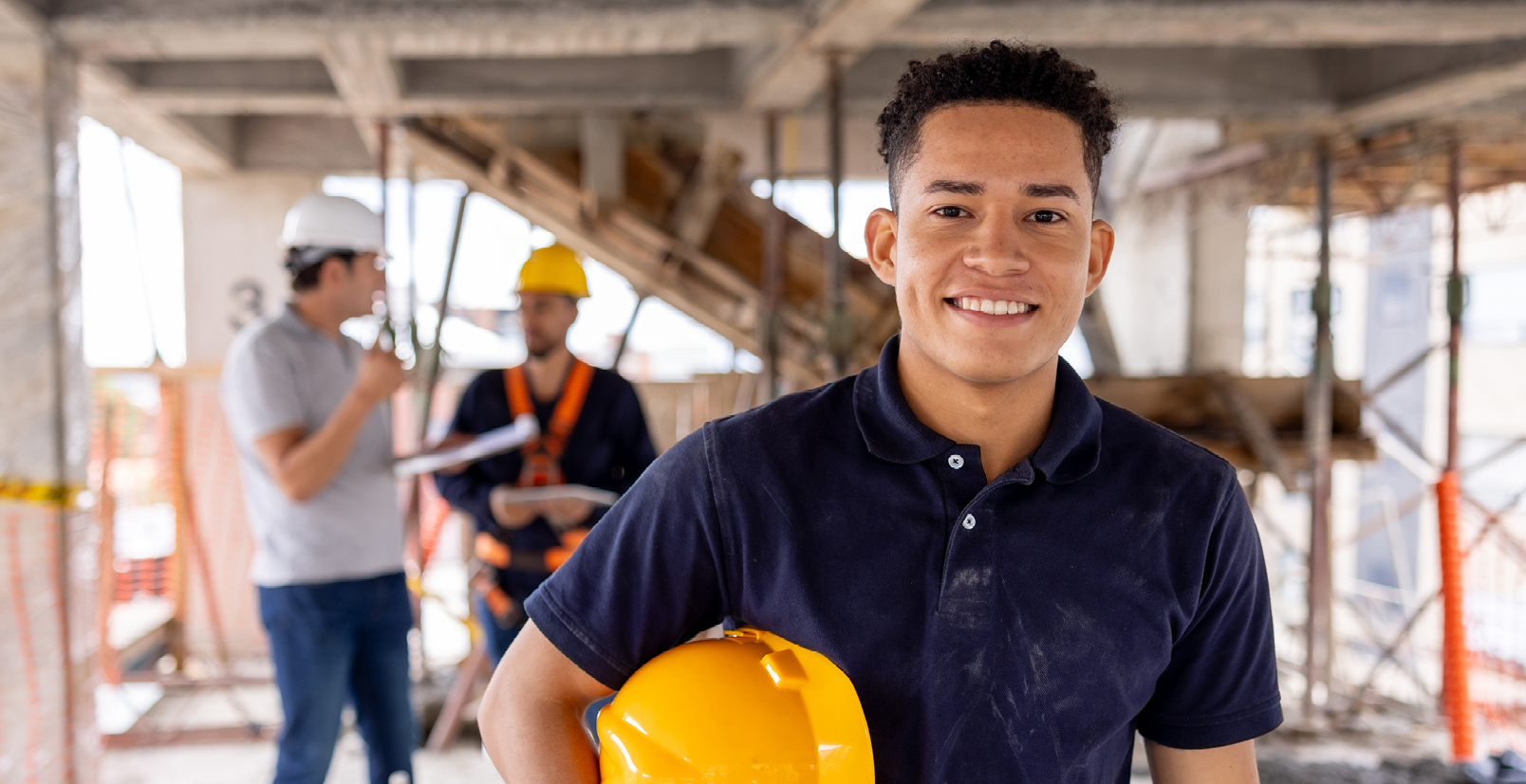 Young man wearing a navy coloured shirt holding his yellow construction site safety hat. He's smiling and facing the camera. There are two males standing in the background having a conversation. The setting is a building site.